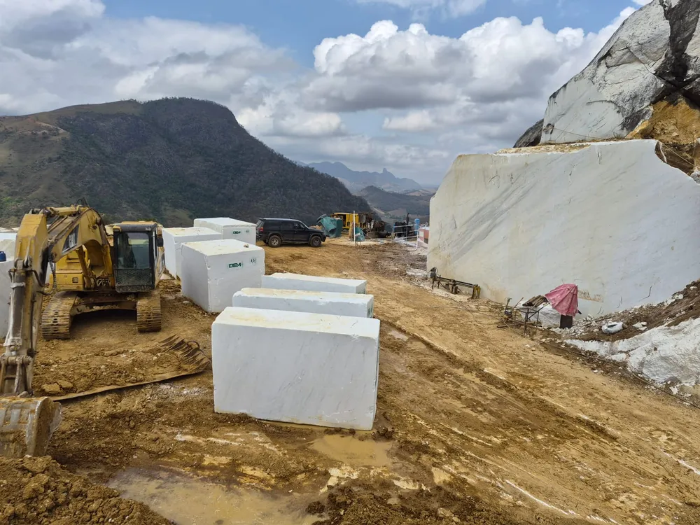 Escavadeira e blocos de mármore identificados com a marca DCM no pátio da jazida DCosta, com montanhas ao fundo